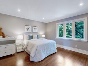 Spacious bedroom with hardwood flooring, recessed lighting, and large windows overlooking mature trees.